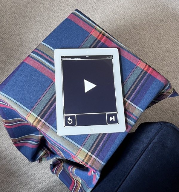 Ruth's iPad on a tartan blanket on the arm of her chair, showing the player: one large play button and two small ones