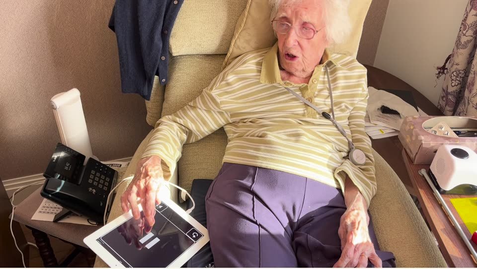 Ruth, 101, in her armchair, with her hand on the iPad beside her that shows her audiobook player
